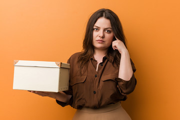 Wall Mural - Young curvy plus size woman holding a box pointing his temple with finger, thinking, focused on a task.