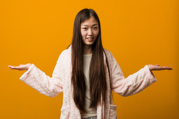 Young chinese woman in pajama makes scale with arms, feels happy and confident.