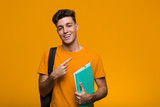 Fototapeta  - Young student man holding books pointing with finger at you as if inviting come closer.