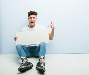 Young man holding a placard sitting on his home floor smiling cheerfully pointing with forefinger away.