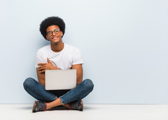 Wall Mural - Young black man sitting on the floor with a laptop smiling confident and crossing arms, looking up