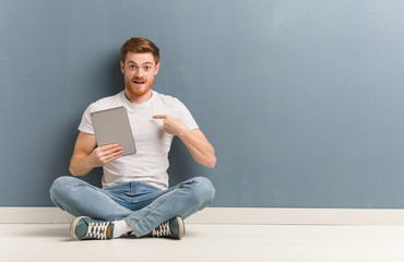 Young redhead student man sitting on the floor surprised, feels successful and prosperous. He is holding a tablet.