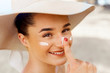 © verona_studio - Beautiful Young woman with sun cream on face.  Female in hat applying  moisturizing lotion on skin.Skin care. Sun protection. Suntan