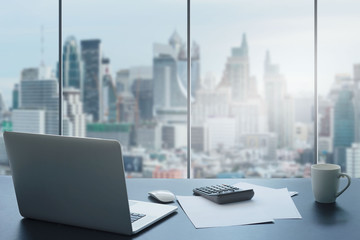 Laptop on table in office room on window city background.Computer and white paper,calculator with a coffee cup on a black desk