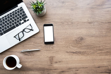 Wood office desk table with smartphone with blank mock up screen, laptop computer, cup of coffee and supplies. Top view with copy space, flat lay.