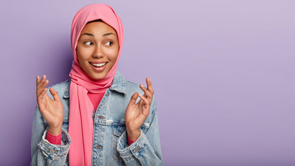 Canvas Print - Smiling delighted Muslim woman clasps hands, looks gladfully aside, has dark skin, wears pink hijab and denim jacket, poses against purple background with blank space for information. Wow, how nice