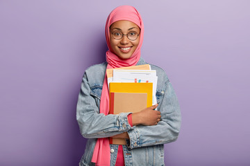 Canvas Print - Beautiful young Arabian woman with dark skin, wears transparent spectacles, holds papers and notepad, has toothy smile, has religious views, studies at college, isolated against purple background