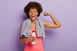 © wayhome.studio - Horizontal shot of happy Afro woman shows biceps, satisfied after active training, holds bottle of water, enjoys healthy lifestyle, isolated over purple background. Sport and exercising concept