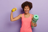 Funny happy dark skinned woman raises hand with dumbbell, shows biceps, holds rolled fitness mat, smiles broadly, wears pink casual vest, isolated over purple background. Fitness training at home