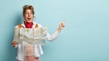 Positive Female Traveler Holds Paper Map, Shows Direction, Being Lost In Unknown Place, Wears Round Spectacles, Red Bandana, Points At Free Space, Has Glad Look, Isolated Over Blue Background