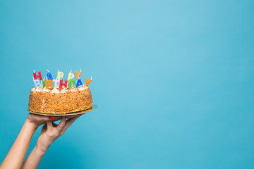 Hands holding a birthday cake with candles and the inscription birthday on a blue background. Copy Space