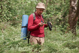Close up portrait of concentrated eldery male wearing red casual sweater, pants and cap, standing in forest with compass and binoculars in hands, tries to find way, backpacking while having vacation.