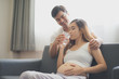 © linghaa - Young pregnant Asian woman is sitting on sofa with her husband are drinking a water near window which felling happy at family's home. To be new Mother and Father, pregnant woman concept.