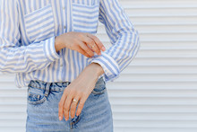 Stylish Woman Wearing Blue Striped Shirt Rolls Up Sleeves Against White Wall. Details Of Trendy Casual Outfit. Street Fashion. Gold Rings On Female Fingers. Jewelry, Accessories, Close Up, Women Hands