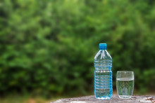 A Bottle Of Water And A Glass Filled With Water On The Surface Of The Stump On The The Green Blurred Foliage Background