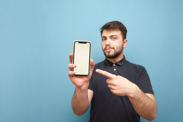 Smiling man holds a smartphone in his hands, and shows his finger on a white screen on a blue background. Bearded man wears a dark T-shirt and shows a smartphone with a white screen in the camera.