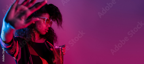 Annoyed fashion young african girl black woman wear stylish pink glasses hold drink say no refuse with stop hand gesture isolated on party purple studio background, banner website design, copy space