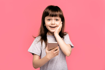 Portrait of a shocked young little child girl looking at mobile phone isolated over pink background. Overjoyed happy excited kid.  rejoices good news, stares at mobile phone, gestures actively 