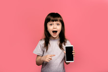 Portrait Of Little Child Girl Over Pink Background Holding Smart Sell Phone On Hand, Screaming With Opened Mouth And Crazy Expression. Surprised Or Shocked Face