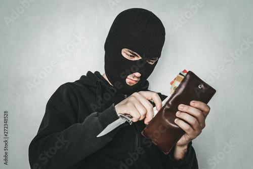 Fotografía Masked criminal at the black background grins looking at the viewer and showing a purse in hand