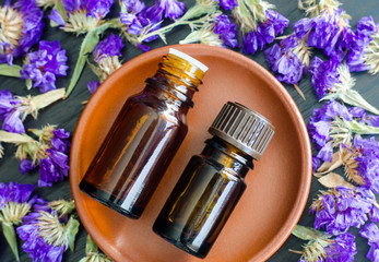 Two small bottles with essential oils on the small ceramic plate. Dark wooden background with dry purple flowers. Aromatherapy, spa and herbal medicine concept. Copy space, top view. 