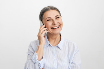 Wall Mural - Studio shot of cheerful ecstatic senior elderly woman wearing elegant blue shirt laughing at good funny joke, showing her perfect straight teeth, enjoying nice phone conversation using mobile