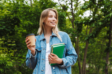 Canvas Print - Photo of cute blonde woman drinking coffee and holding books while walking in green park