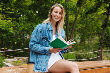 Canvas Print - Photo of cute happy woman wearing reading book and drinking coffee while sitting on bench in green park