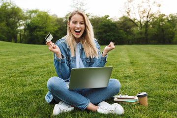 Poster - Photo of adorable cheerful woman holding credit card and using laptop for purchase while sitting on green grass in park