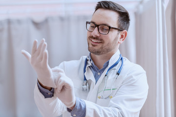 Young smiling Caucasian male doctor in white uniform, with eyeglasses and stethoscope around neck putting rubber gloves before patient examination.