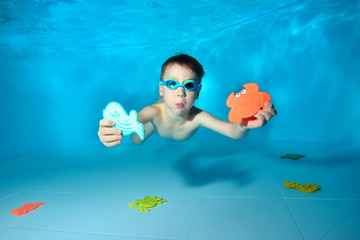 Cute sports baby swims underwater and holds sea toys in his hand. He looks at the camera and smiles. Portrait. Underwater photography. Horizontal orientation