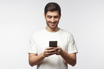 Wall Mural - Studio portrait of young european caucasian man isolated on gray background wearing white t-shirt standing in front of camera, looking attentively with smile at screen of smartphone he is holding