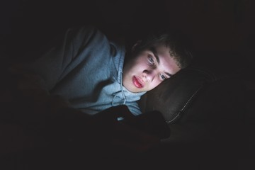 Wall Mural - Teenager lying down on a couch in the dark. The light from the screen of his smartphone is illuminating his face.