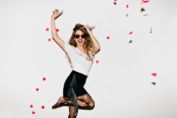 Emotional female model in pantyhose having fun in studio. Funny girl in sunglasses looking at heart confetti.