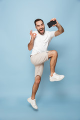 Wall Mural - Full length photo of young caucasian man in casual white t-shirt screaming while jumping and taking selfie on smartphone