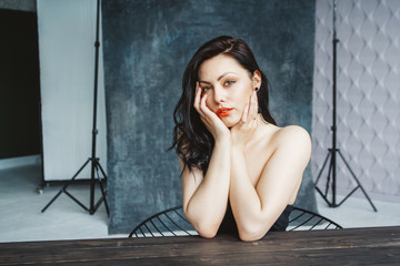 Portrait of young beautiful brunette woman sitting at table on dark gray copy space background. Gorgeous natural beauty girl in cute dress indoors. A look at the camera