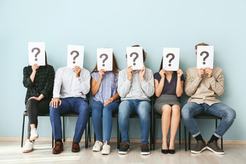 Canvas Print - Young people hiding faces behind paper sheets with question marks while waiting for job interview indoors