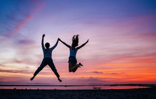 Two Running And Jumping Girlfriends On The Sunset Sea Beach. Hand In Hand Concept Image.