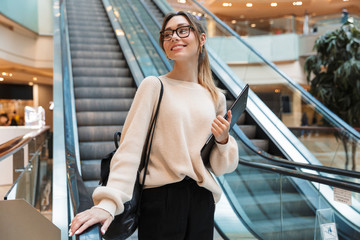 Sticker - Photo of cute young woman smiling and holding clipboard while going down escalator in building indoors