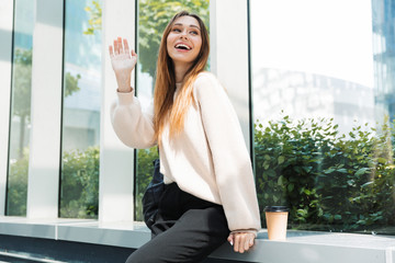 Sticker - Photo of caucasian charming woman smiling and waving her hand while sitting near window in building indoors