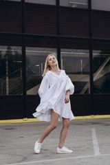 The girl is dressed in a stylish white, lush summer dress, dynamically poses for the photo, posing against street , urban clothing style. Street photography