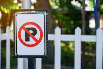 White red and black no parking sign on the background of a white fence and green leaves.