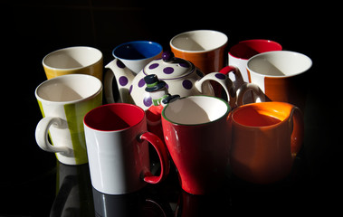 Multiple colorful ceramic mugs and a tea pot shot from aside on a black background