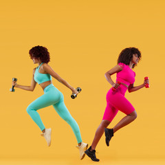 Wall Mural - Beautiful African American and caucasian women running. Two strong athletic, women sprinter or runner on yellow background with dumbbells wearing color sportswear. Fitness and sport motivation.