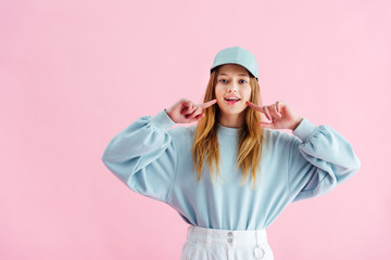 Wall Mural - pretty teenage girl in cap pointing with fingers at smile isolated on pink