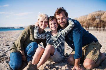 Wall Mural - Young family with small boy sitting outdoors on beach, looking at camera.