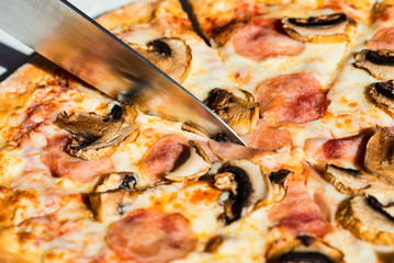 Close up knife slicing pizza with salami, mushrooms and cheese close up