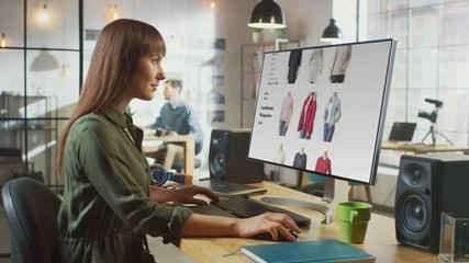 Poster - Beautiful Brunette Female Scrolls Through Online Fashion Store on Her Personal Computer with Big Display. She Works in a Cool Office Loft. Other Male Creative Colleague Walks in the Background.