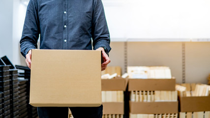 Young courier man carrying cardboard box near product shelves in warehouse. Parcel shipping service or delivery packaging concept