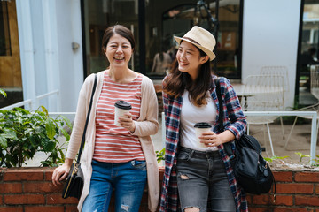 group of young asian girls sitting on garden outdoors cafe bar holding paper coffee drink. two laughing women smiling having fun chatting on sunny day. modern cozy restaurant shop in background.